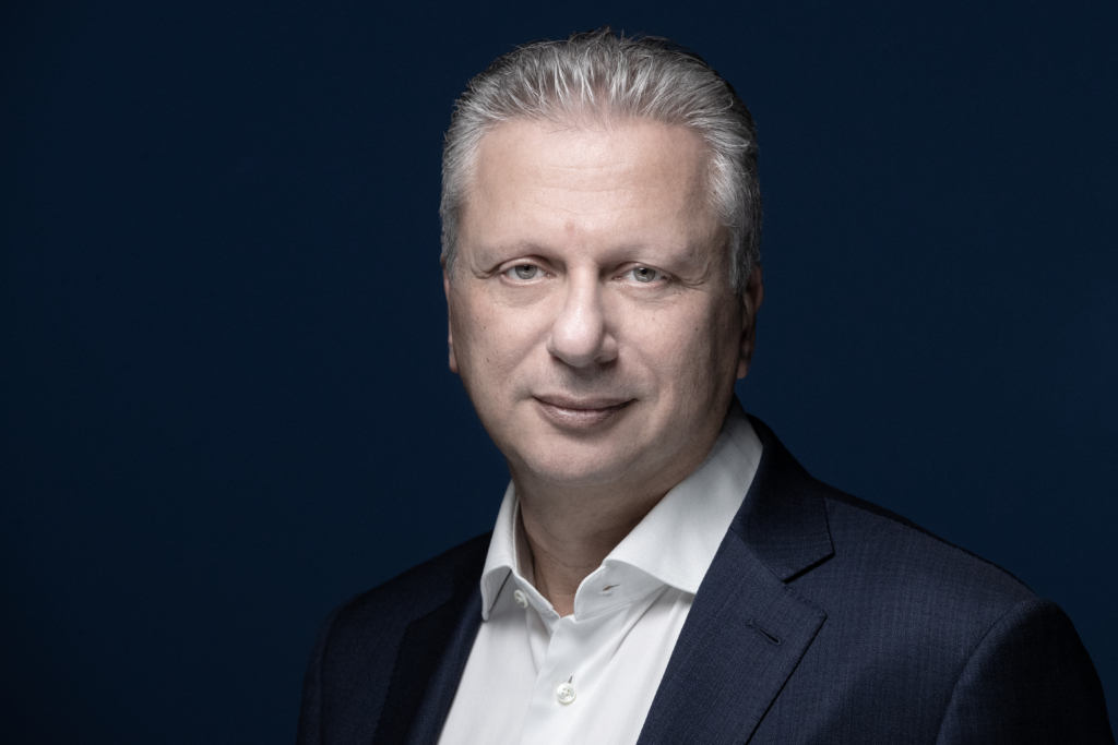 Aiman Ezzat, CEO of Capgemini, looks to camera. He wears a deep navy suit.